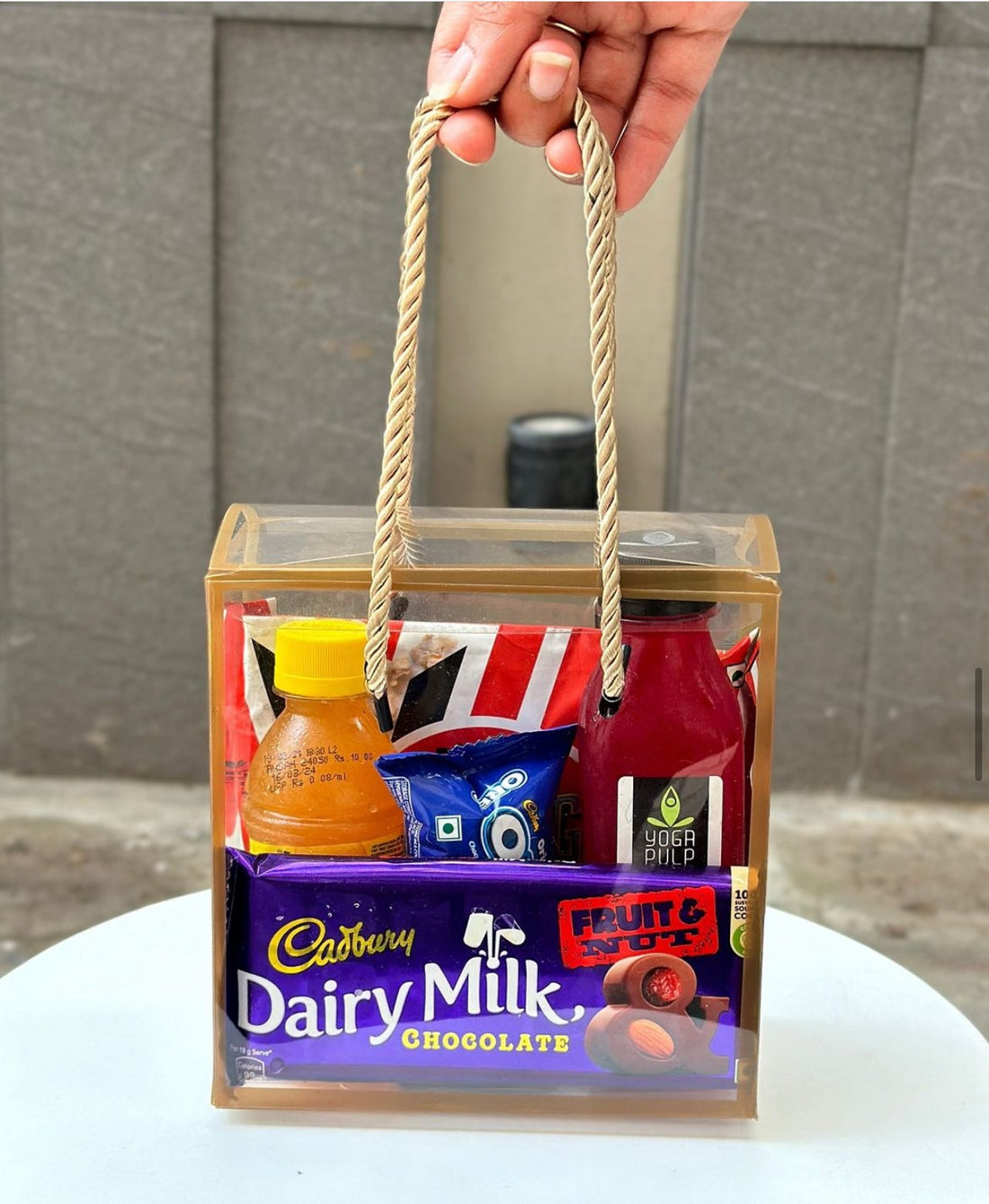 Hand holding a gift basket with Cadbury Dairy Milk chocolate and other items against a neutral background.